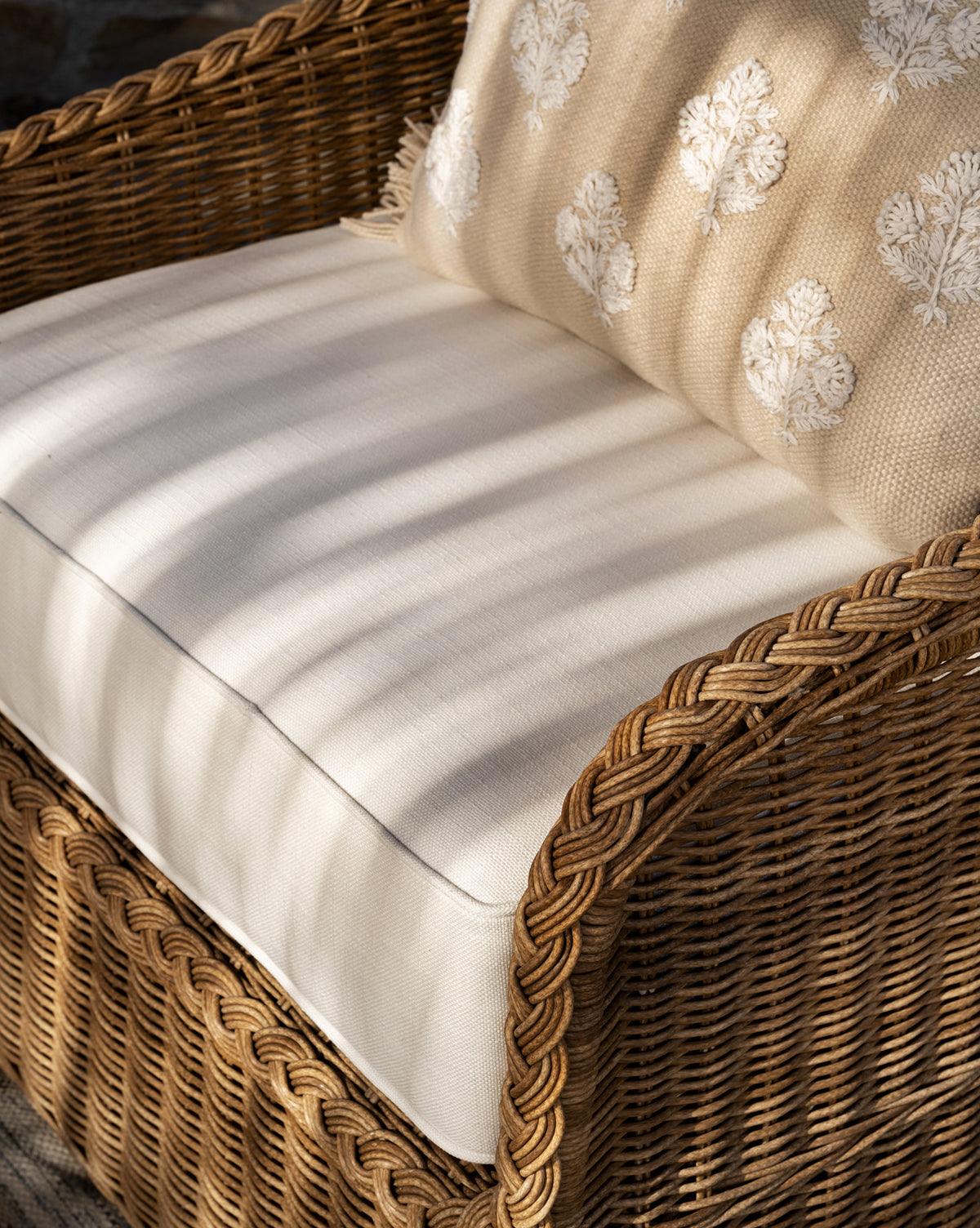 A close-up of the Haviland Outdoor Lounge Chair, crafted from weather-resistant wicker with a cream cushion and a beige pillow adorned with white floral patterns, illuminated by gentle sunlight and shadow by McGee & Co.
