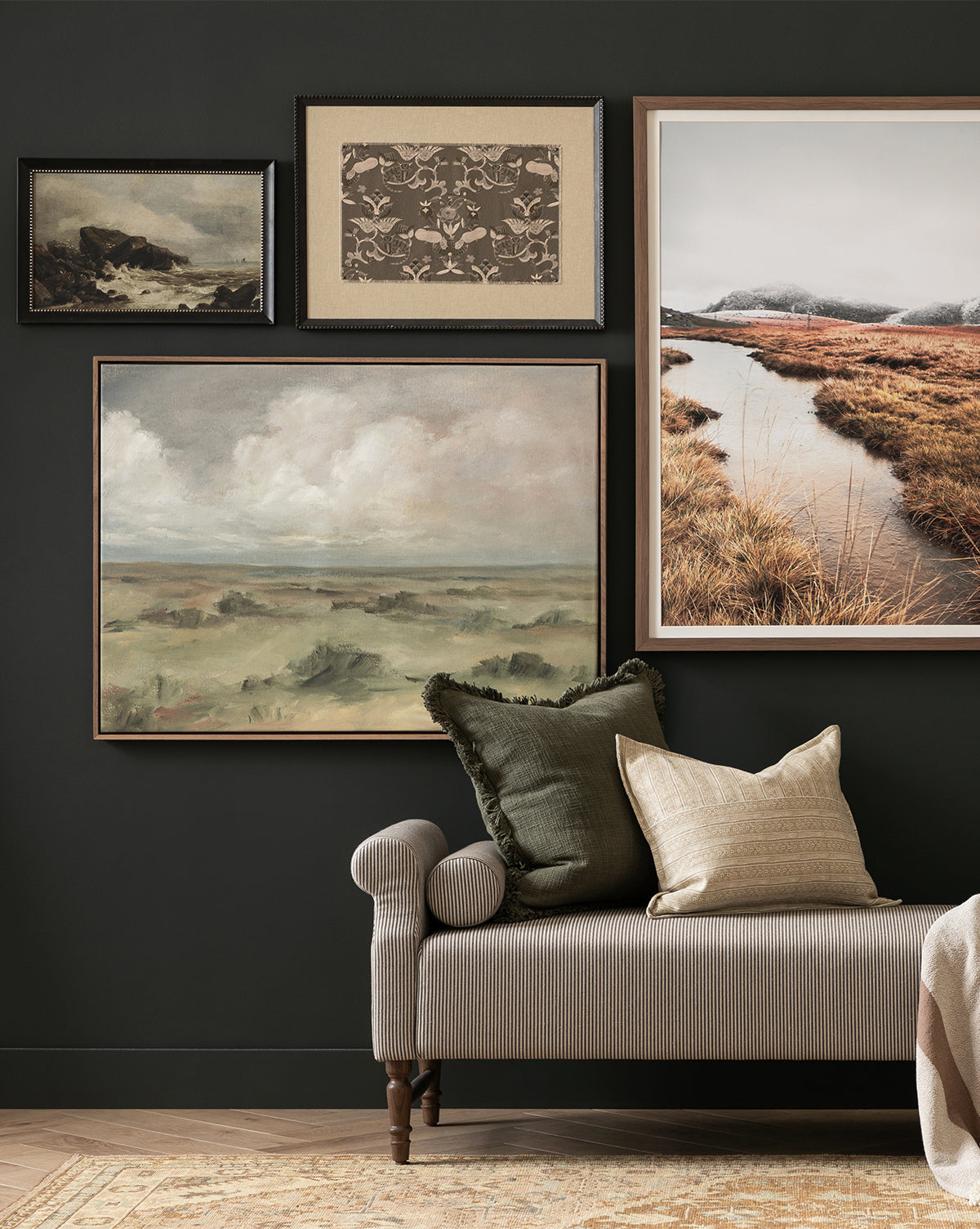 A cozy room features the Fable striped chaise lounge with dark green and beige pillows, set beneath a gallery wall showcasing four black-framed landscape and nature artworks, including a striking textural piece on the dark wall, by McGee & Co.
