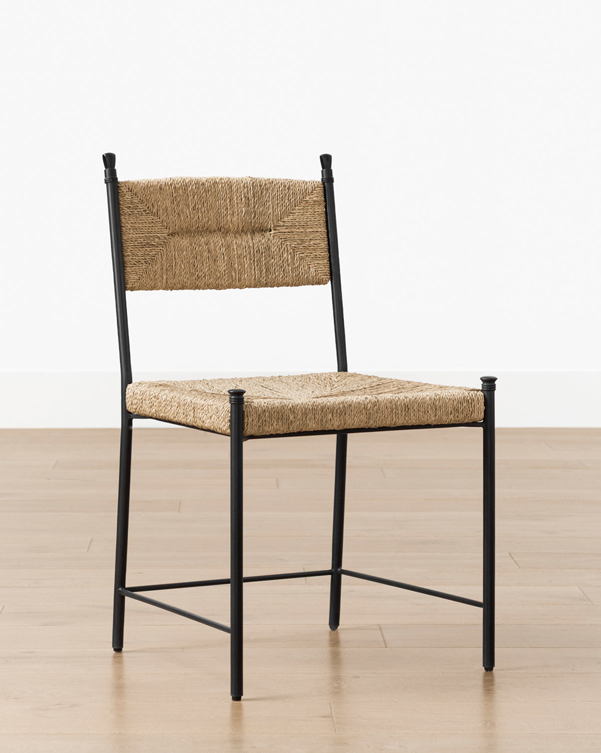 The Peregrine Chair features a black metal frame with woven natural fiber seat and backrest, shown on a light wood floor against a white wall. Ideal as a stylish dining chair or accent piece in any modern space by McGee & Co.