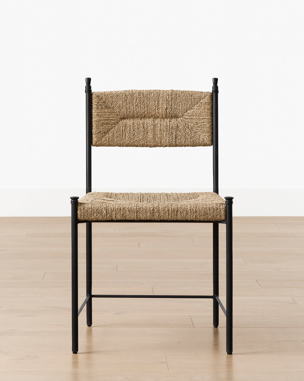 A Peregrine Chair featuring a black metal frame and woven natural fiber seat and backrest sits on a light wood floor before a plain white wall, by McGee & Co.