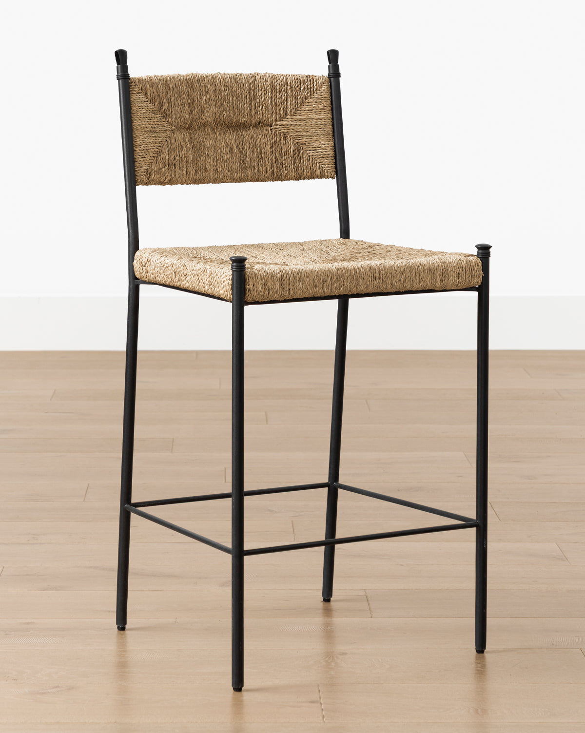 The Peregrine Stool by Makerspalm features a black steel frame with woven natural rattan seat and backrest, set on a light wooden floor against a plain white wall. McGee & Co.