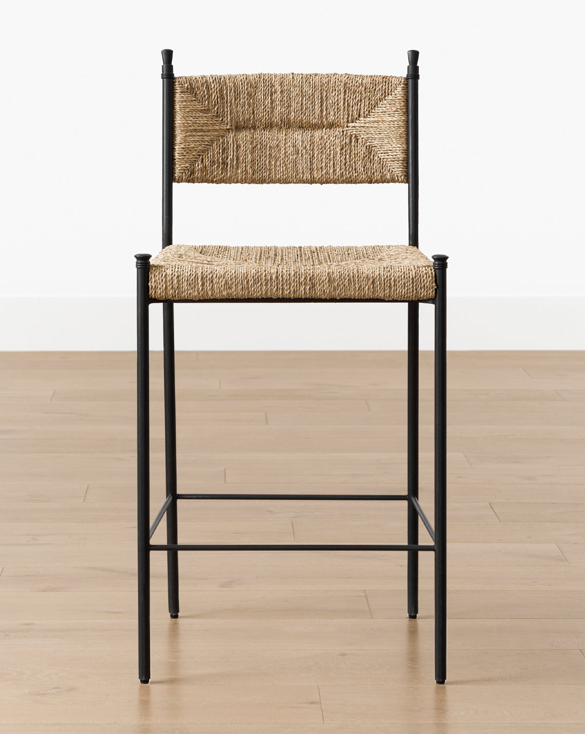 The Makerspalm Peregrine Stool, featuring a black metal frame with woven seagrass seat and backrest, sits on a light wood floor against a white wall—perfect as a stylish kitchen dining stool. McGee & Co.