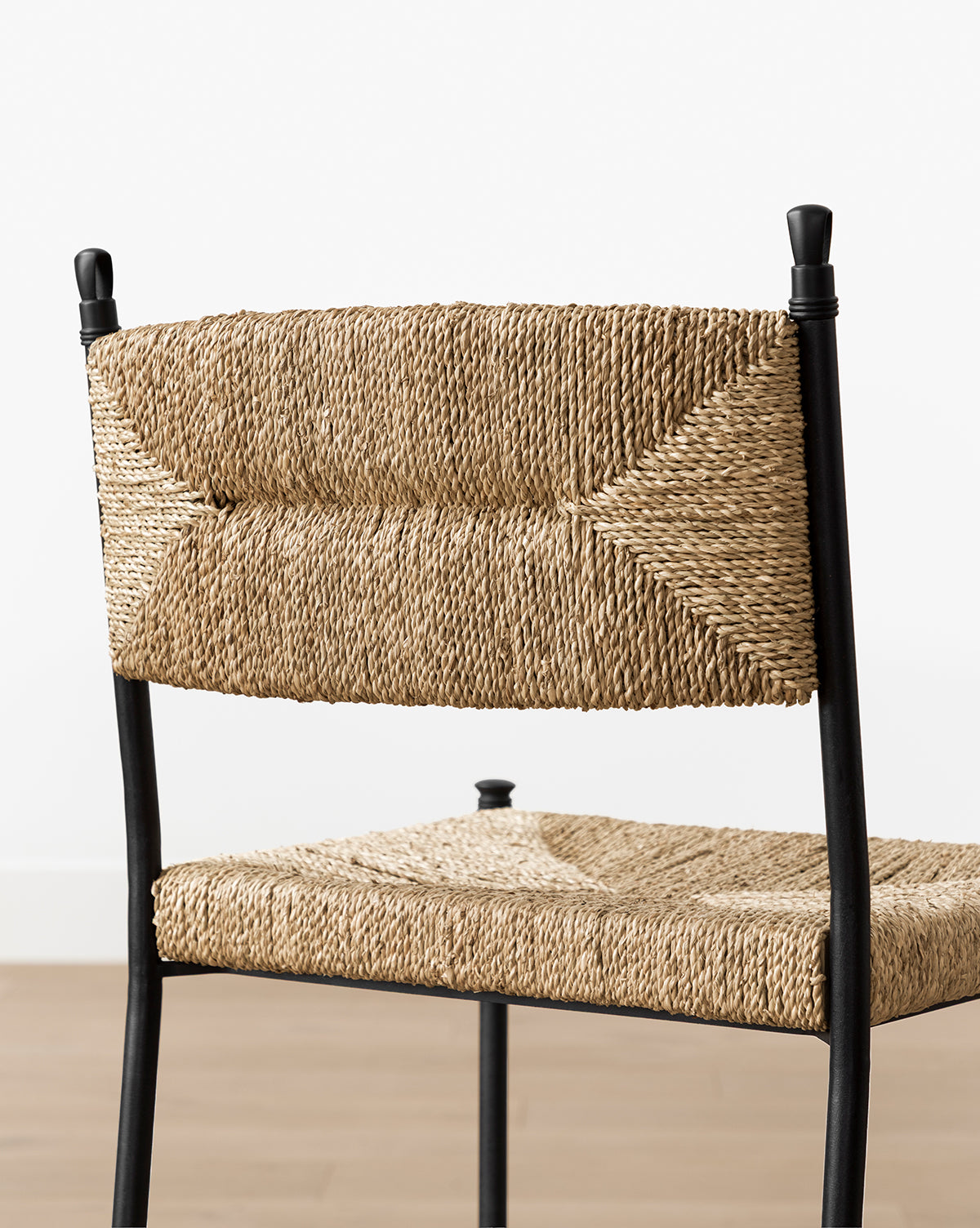 The Peregrine Stool by Makerspalm features a black steel frame with woven seagrass seat and backrest in natural fibers, shown against a plain white wall and wooden floor. McGee & Co.