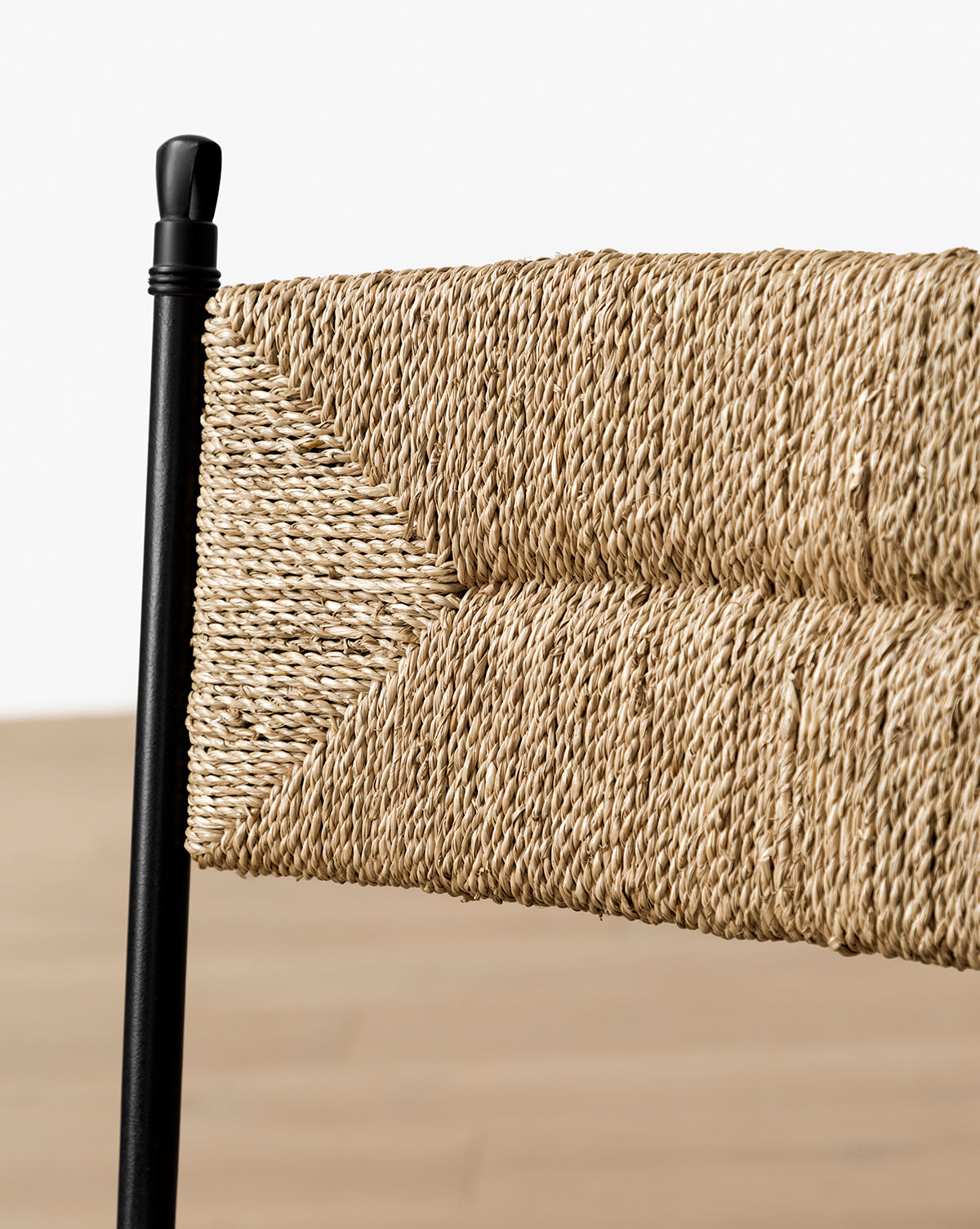 Close-up of the Peregrine Stool by Makerspalm—featuring a chevron-patterned woven seagrass and natural rope backrest, black steel frame, and blurred light wood floor background—ideal as a stylish kitchen dining stool. McGee & Co.