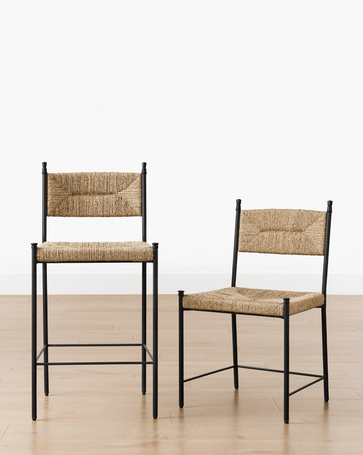 Two Peregrine Stools by Makerspalm, featuring black steel frames and woven straw seats and backs, are displayed on a light wood floor against a plain white background. One stool is taller with a higher seat and backrest than the other. McGee & Co.