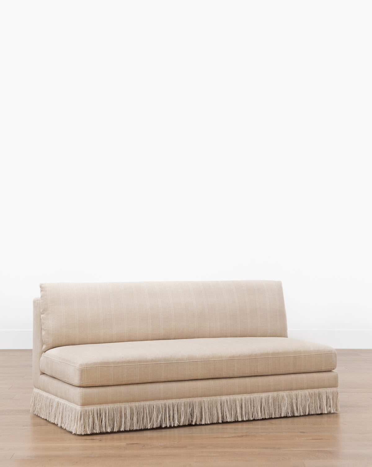 The Philippa Settee (Ready to Ship) is a beige, armless linen seat with subtle vertical stripes, a low backrest, a seat cushion, and decorative fringe along the bottom edge, shown on a wooden floor against a white background by McGee & Co.