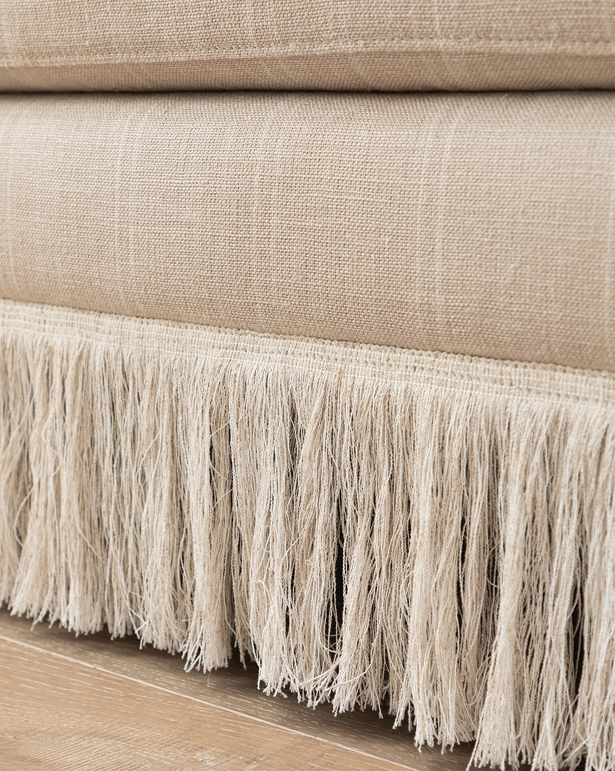 The Philippa Settee (Ready to Ship) features beige woven fabric with visible texture and a white fringe detail at the bottom, adding a stylish touch above a wooden floor, by McGee & Co. The soft, slightly uneven fringe enhances its modern appeal.