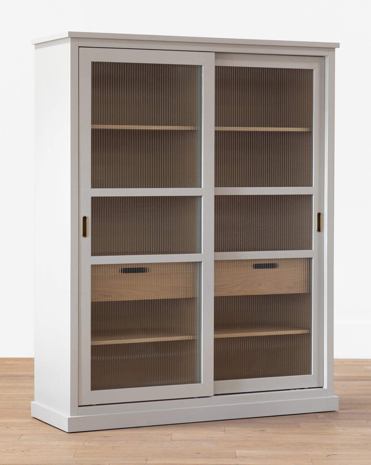 The Phillips Cabinet features a painted oak finish, reeded glass doors with vertical ribbed texture, and wooden shelves inside, by McGee & Co. It stands on a wooden floor against a plain white background.