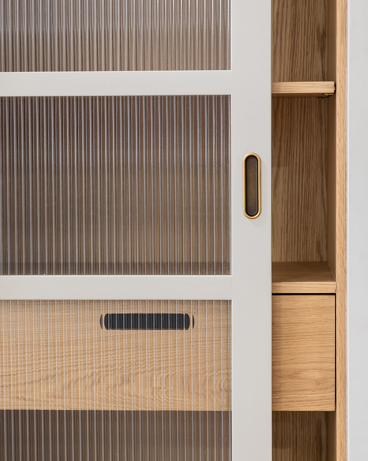 The Phillips Cabinet features a close-up of painted oak with reeded glass doors, recessed metal or antique brass handles, inner shelves and drawers, all in a light wood and white finish, by McGee & Co.
