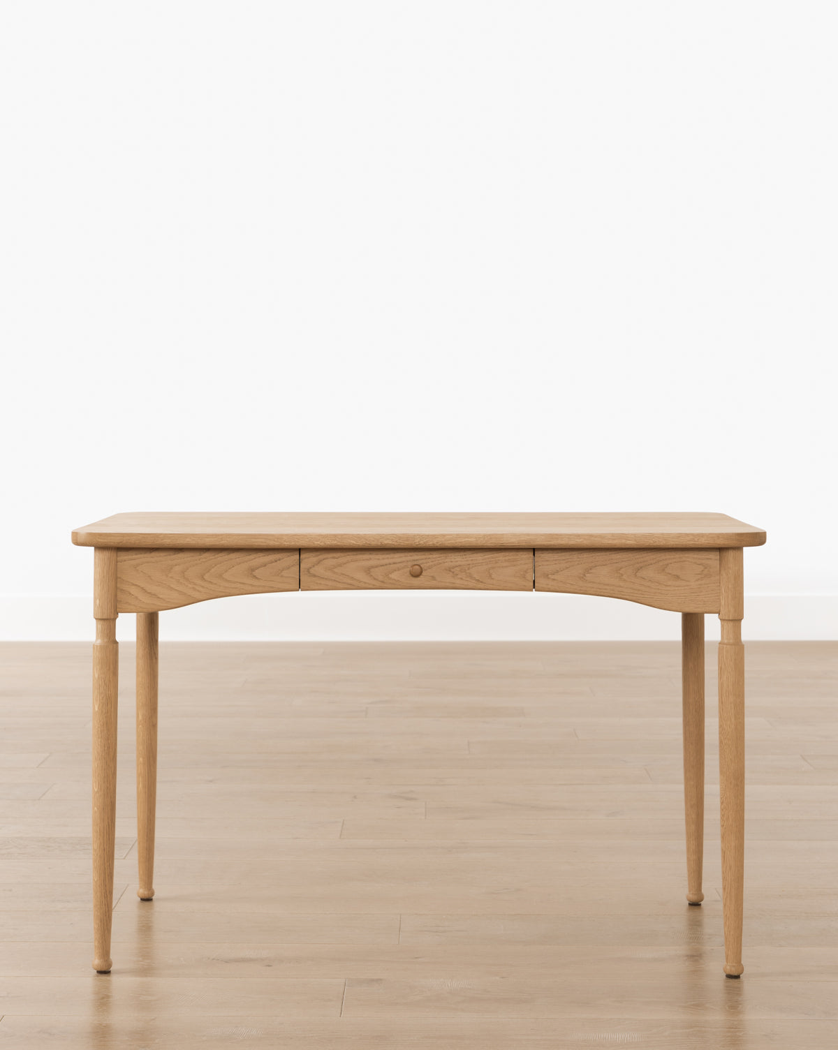 The Phipps Desk features a simple, handcrafted design with four slender legs and a single center drawer, placed on a light wood floor against a plain white wall, by McGee & Co.