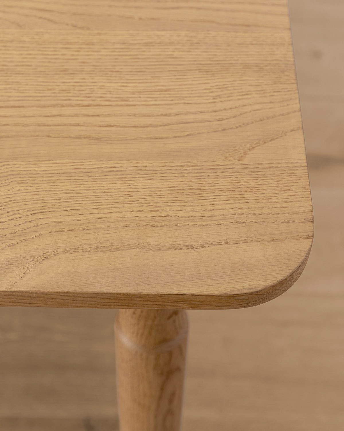 Close-up of the Phipps Dining Table’s handcrafted corner, featuring a smooth, rounded edge and visible grain patterns. The partially visible leg sits atop a similar light wood floor, by McGee & Co.