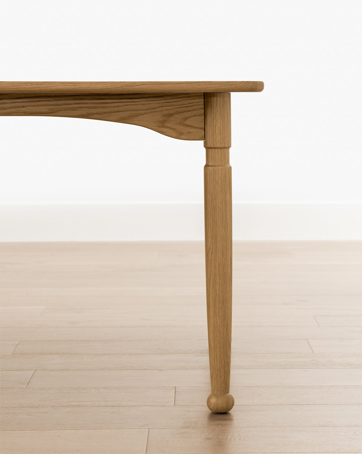 A close-up of the Phipps Dining Table’s handcrafted leg with a natural finish, shown on a light wood floor beside a white wall and simple baseboard, by McGee & Co.