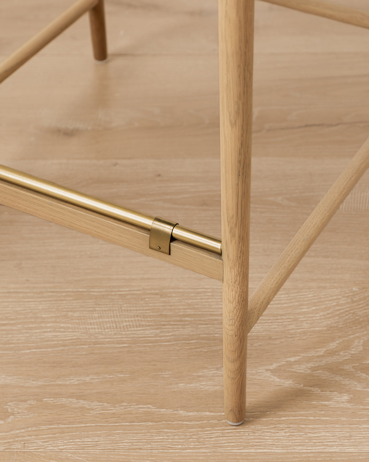 Close-up of a Phipps Woven Bench leg in honey oak with a horizontal brass support bar, standing on a light wood floor. The bench features a woven seagrass seat by McGee & Co.