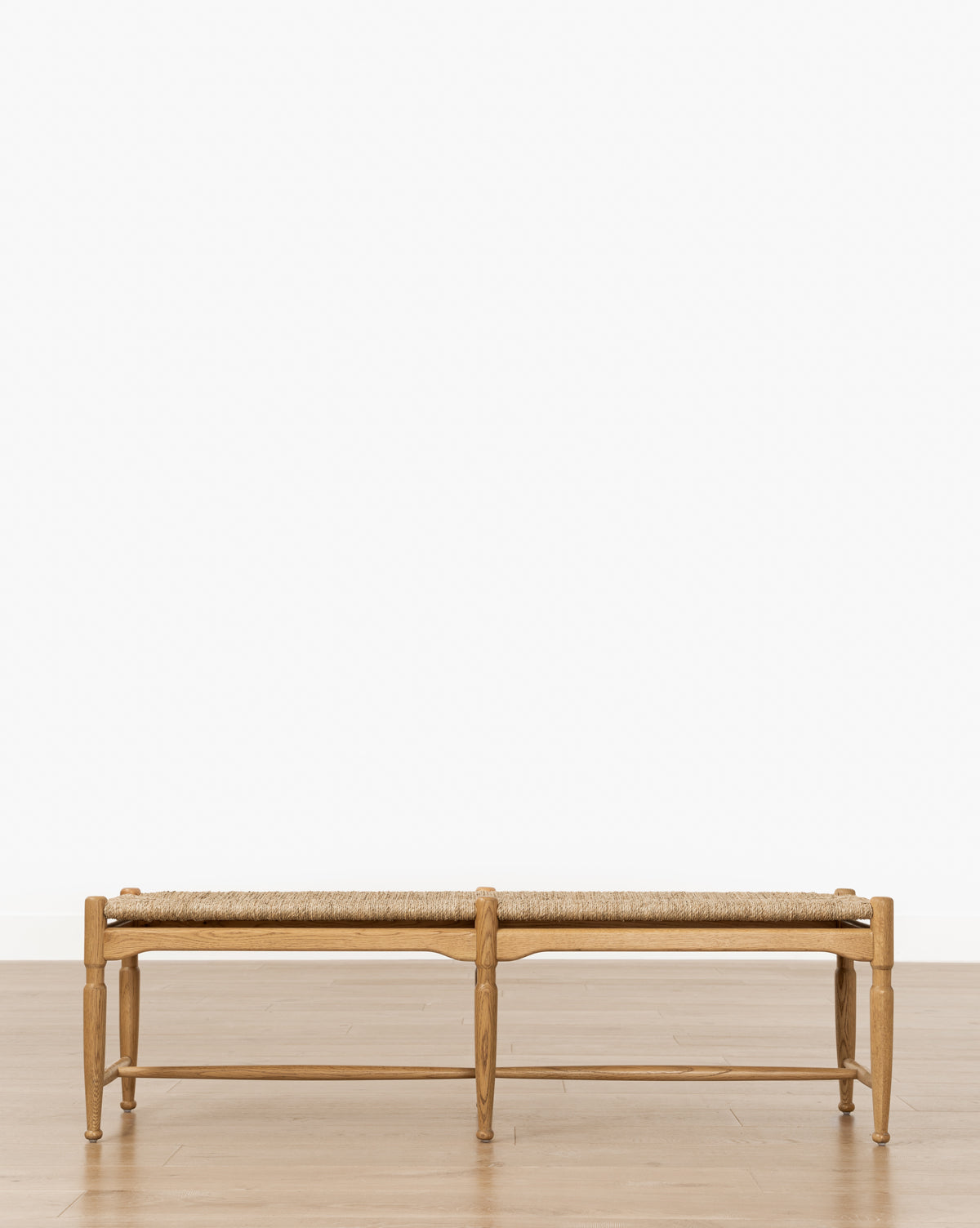 The Phipps Woven Bench, featuring a seagrass seat and wooden frame, is centered on a light honey oak floor against a plain white wall, by McGee & Co.