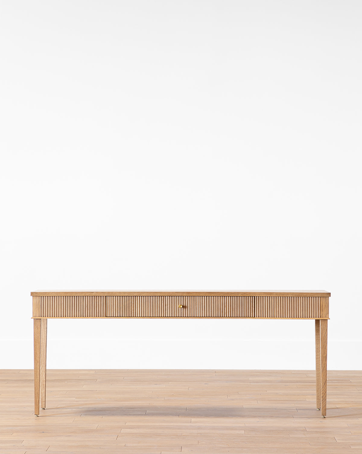 The Pieter Console, a minimalist wooden piece with a concealed drawer, four tapered legs, and subtle reeded detail, stands on a light wood floor against a plain white wall by McGee & Co.