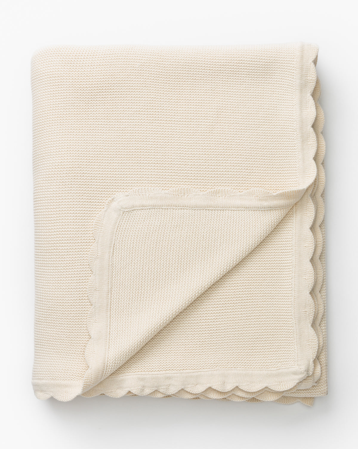 The Pinnacle Knit Kids Blanket, made from 100% cotton in cream with scalloped edges, lies on a white surface. A corner is folded over to reveal the soft texture and edge detail of this cozy heirloom baby blanket.