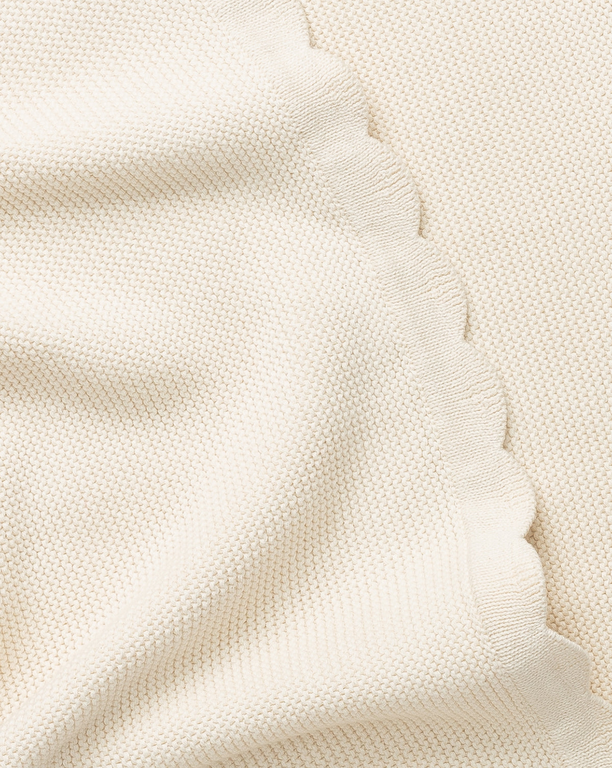 A close-up of the Pinnacle Knit Kids Blanket—100% cotton in cream, with a subtle knit texture and wavy scalloped edge. Softly draped folds make it perfect as an heirloom baby blanket.
