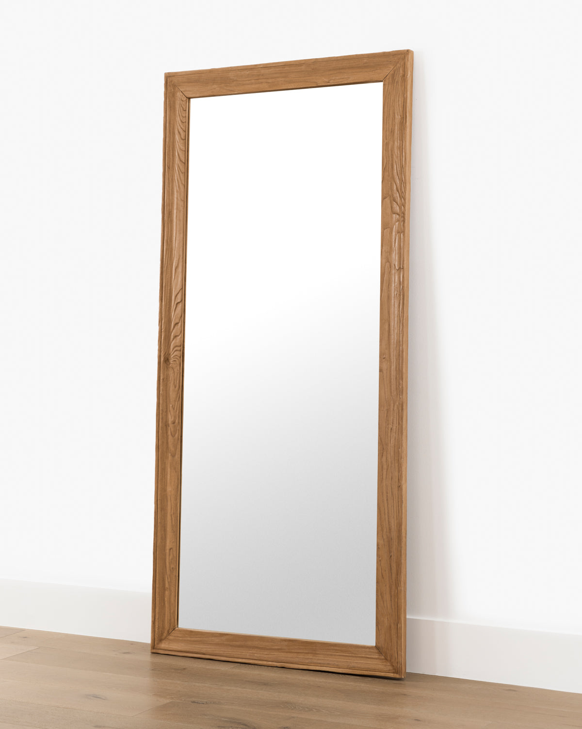 The Pippen Floor Mirror, featuring a large rectangular frame in a honey elm finish, leans against a white wall on a wooden floor, by McGee & Co.