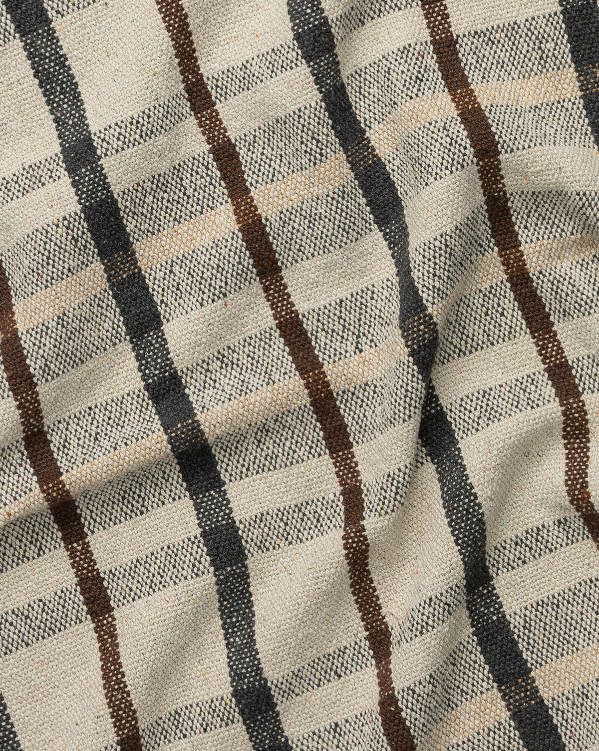A close-up of the Plaid Tasseled Throw shows its beige fabric with a classic plaid pattern in black, brown, and tan stripes. Slightly wrinkled for added texture, it’s finished with braided tassels for timeless style.