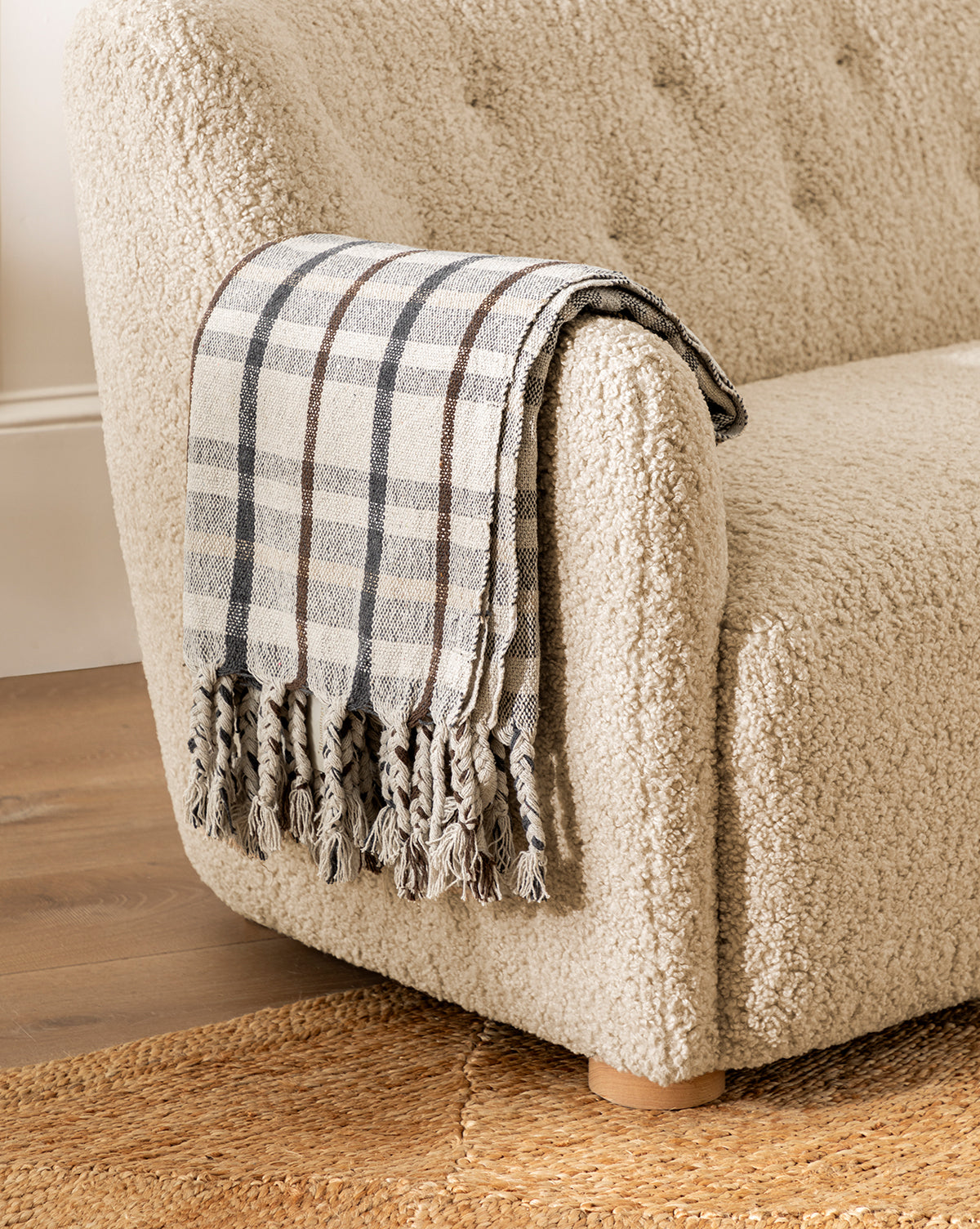 A soft, cream boucle armchair draped with the Plaid Tasseled Throw sits atop a woven jute rug, set against warm wooden flooring in the background.
