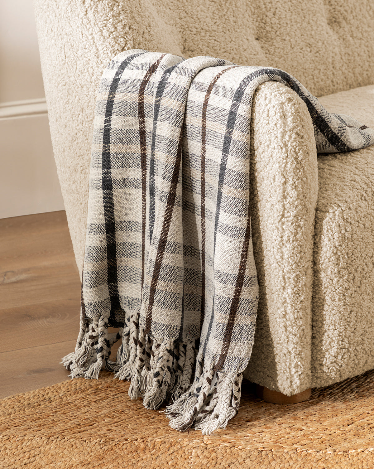A cozy beige armchair with textured fabric features the Plaid Tasseled Throw, showcasing a classic plaid pattern and braided tassels draped over one arm. The chair rests on a woven rug next to a wooden floor.