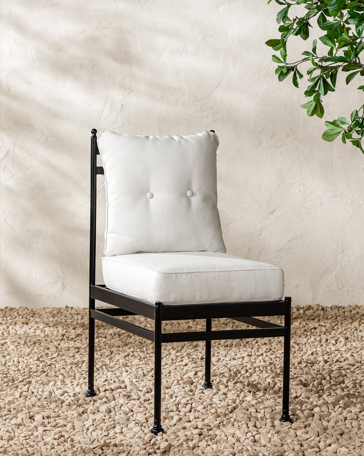 The Pomeroy Outdoor Dining Chair features a black metal frame and white tufted Pasadena White performance fabric cushions. It sits on light pebbles near a leafy green plant against a textured beige wall, by McGee & Co.