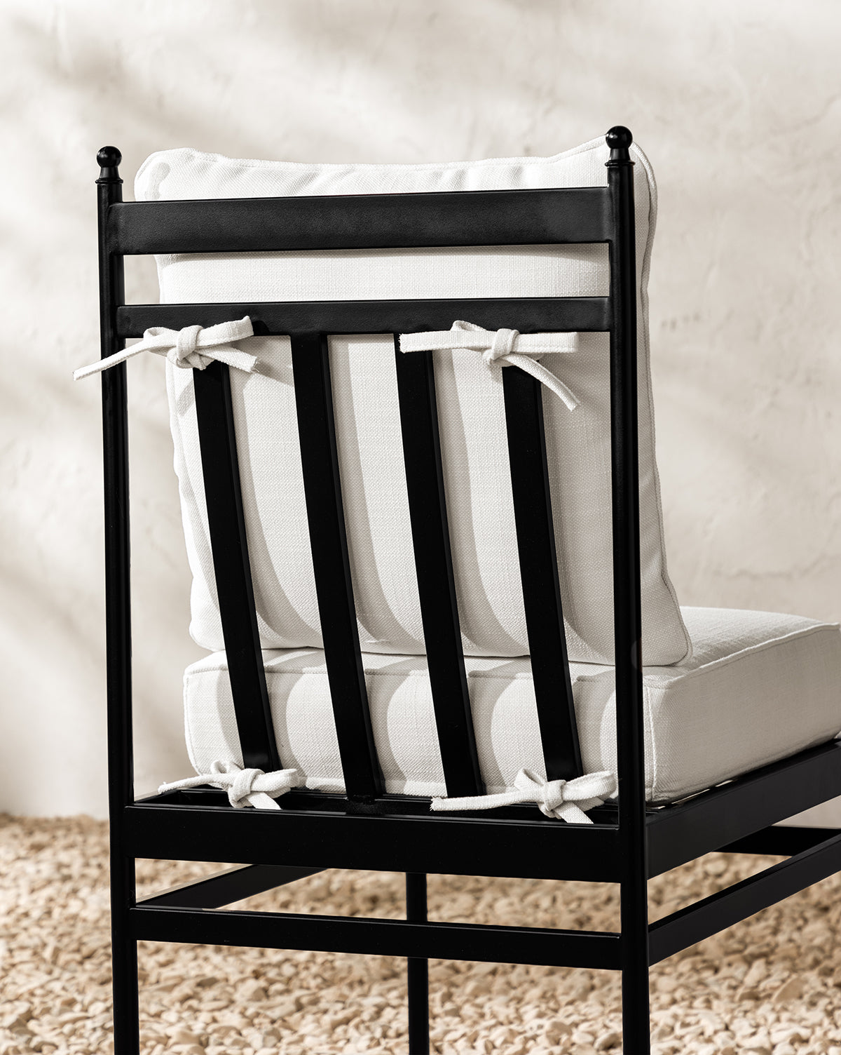 A Pomeroy Outdoor Dining Chair with a black metal frame and Pasadena White cushions secured by white ties stands on light pebbles in front of a textured beige wall, by McGee & Co.