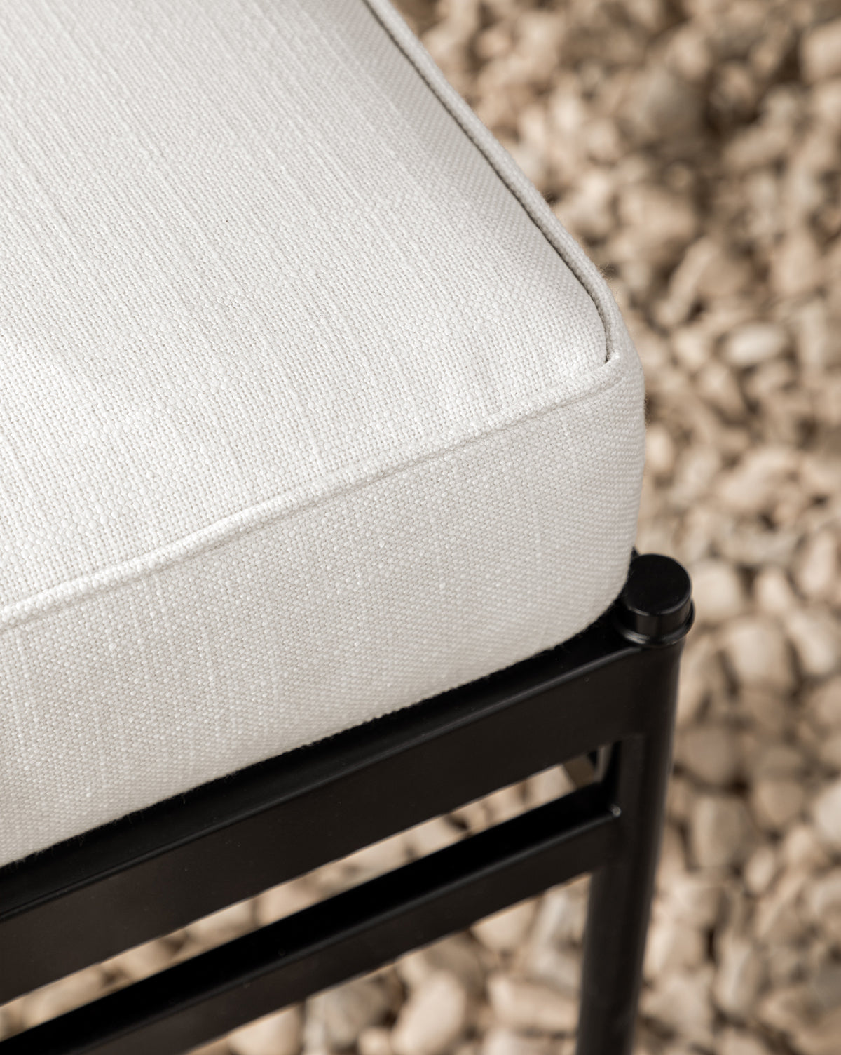 Close-up of a Pasadena White performance fabric cushion on the Pomeroy Outdoor Dining Chair’s black metal frame, with small beige pebbles scattered on the ground in the background, by McGee & Co.