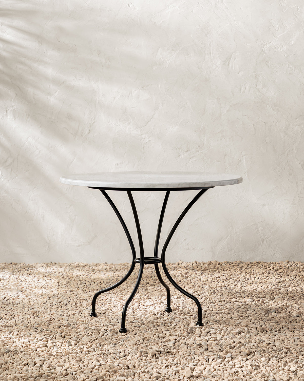 The Pomeroy Outdoor Bistro Table, featuring a marble top and black metal legs, stands on beige pebbles by a textured off-white wall with soft light and subtle shadows—a refined addition to luxury outdoor spaces, by McGee & Co.