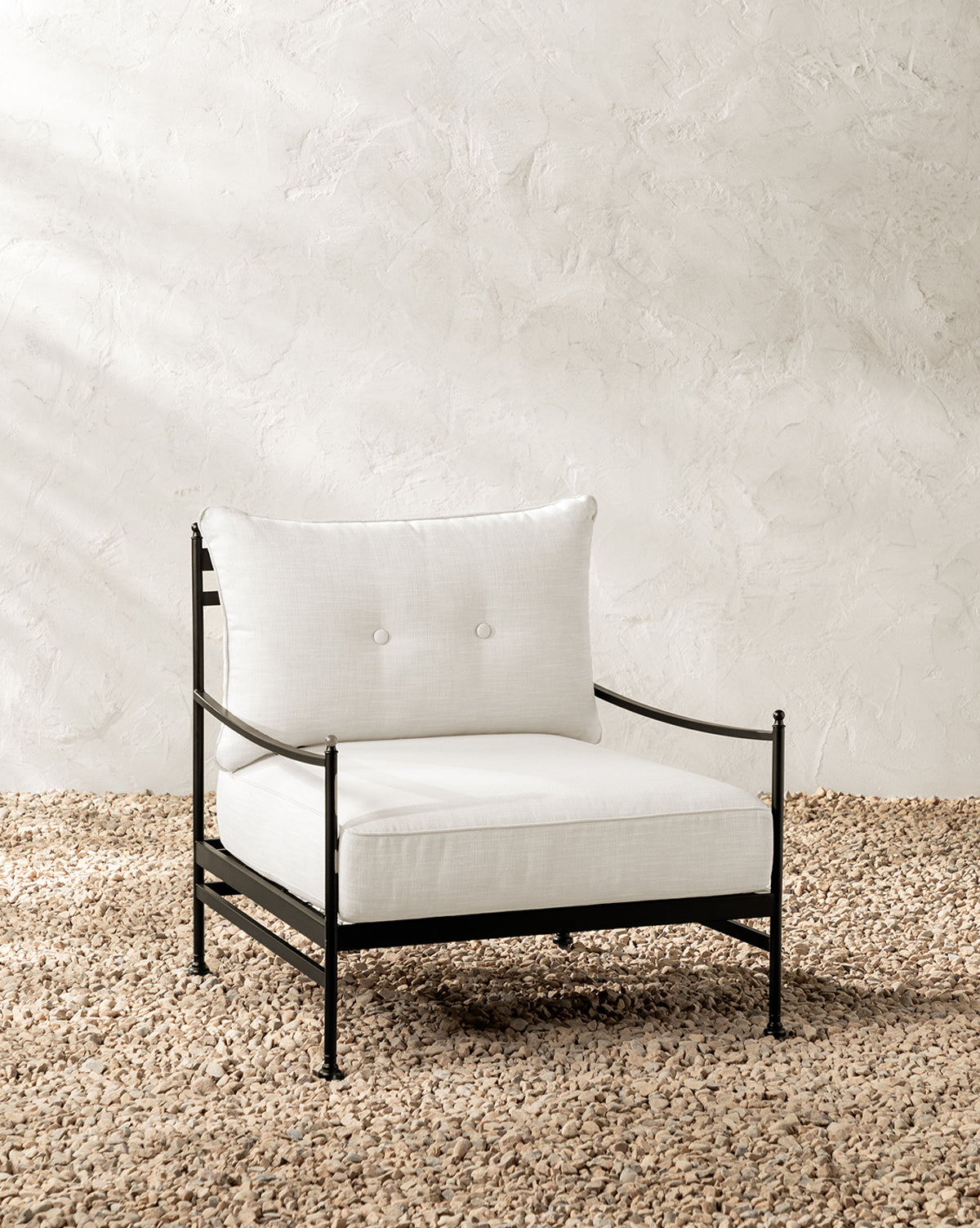 The Pomeroy Outdoor Lounge Chair has a powder-coated steel frame with white outdoor performance fabric cushions, shown on a pebbled surface against a light textured wall, by McGee & Co.