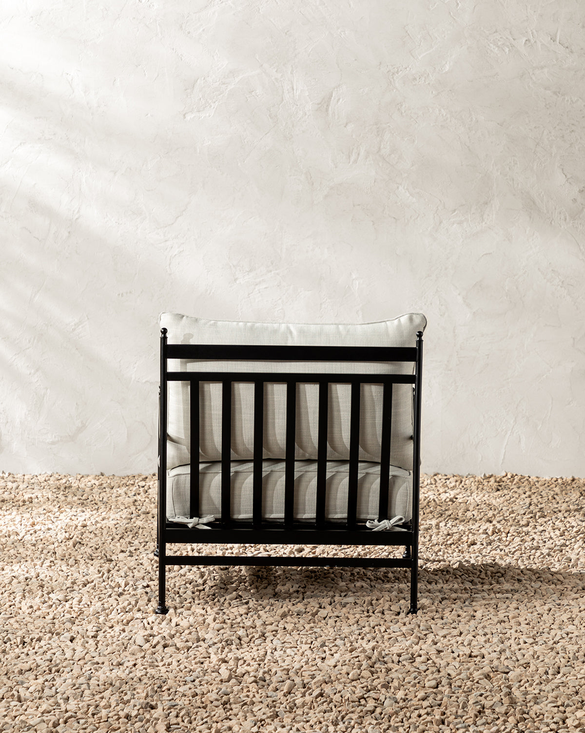 The Pomeroy Outdoor Lounge Chair has a powder-coated steel frame and outdoor performance fabric cushion, shown on beige pebbles with a textured off-white wall backdrop, by McGee & Co.