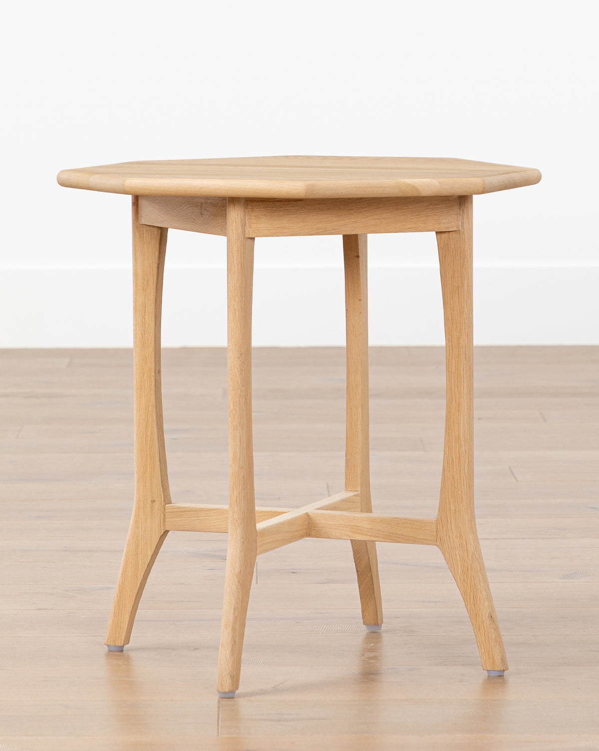 The Posie Oak Side Table features a small, light oak round top and four curved legs. Placed on a wooden floor against a white wall, this accent table adds natural warmth and elegance to any room, by McGee & Co.
