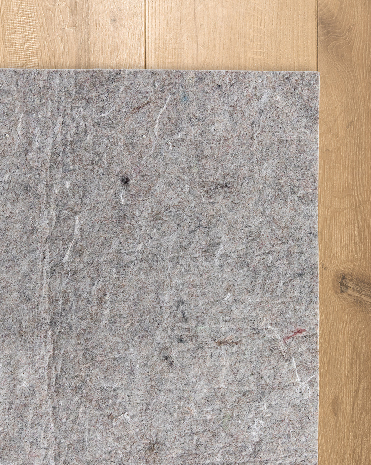 A Premium Indoor Rug Pad lies on a wooden floor, covering part of the surface. Its grey felt features a rough, fibrous texture with visible colored fibers, providing added protection for your floors, by McGee & Co.