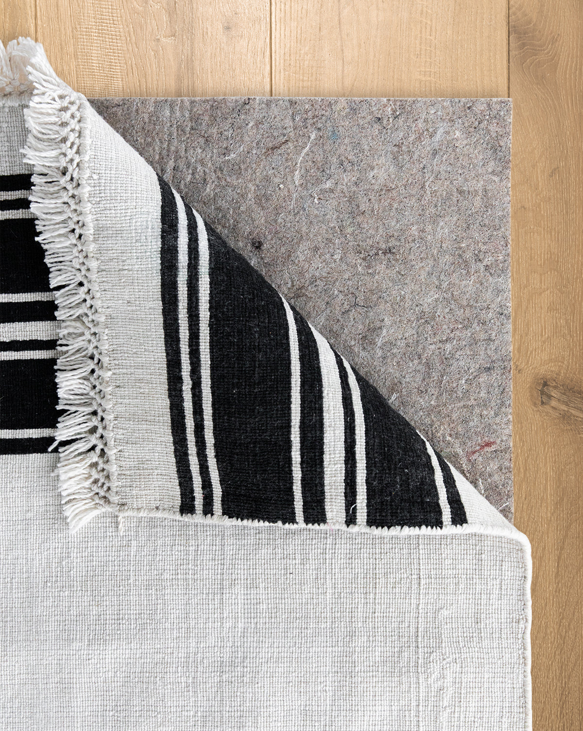 A black and white woven rug with fringed edges is folded at the corner, showing a Premium Indoor Rug Pad with no-slip grip for floor protection on a light wood floor, by McGee & Co.