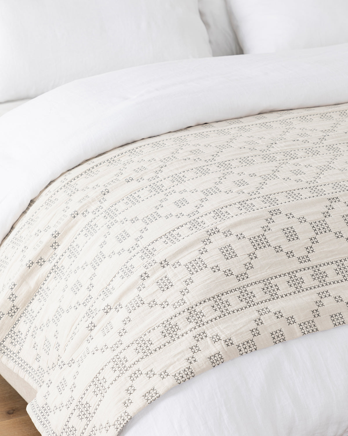 A close-up of a bed with crisp white bedding and the Prescott Coverlet, showcasing a geometric black embroidered pattern, draped across the foot of the bed on a wooden floor, by McGee & Co.