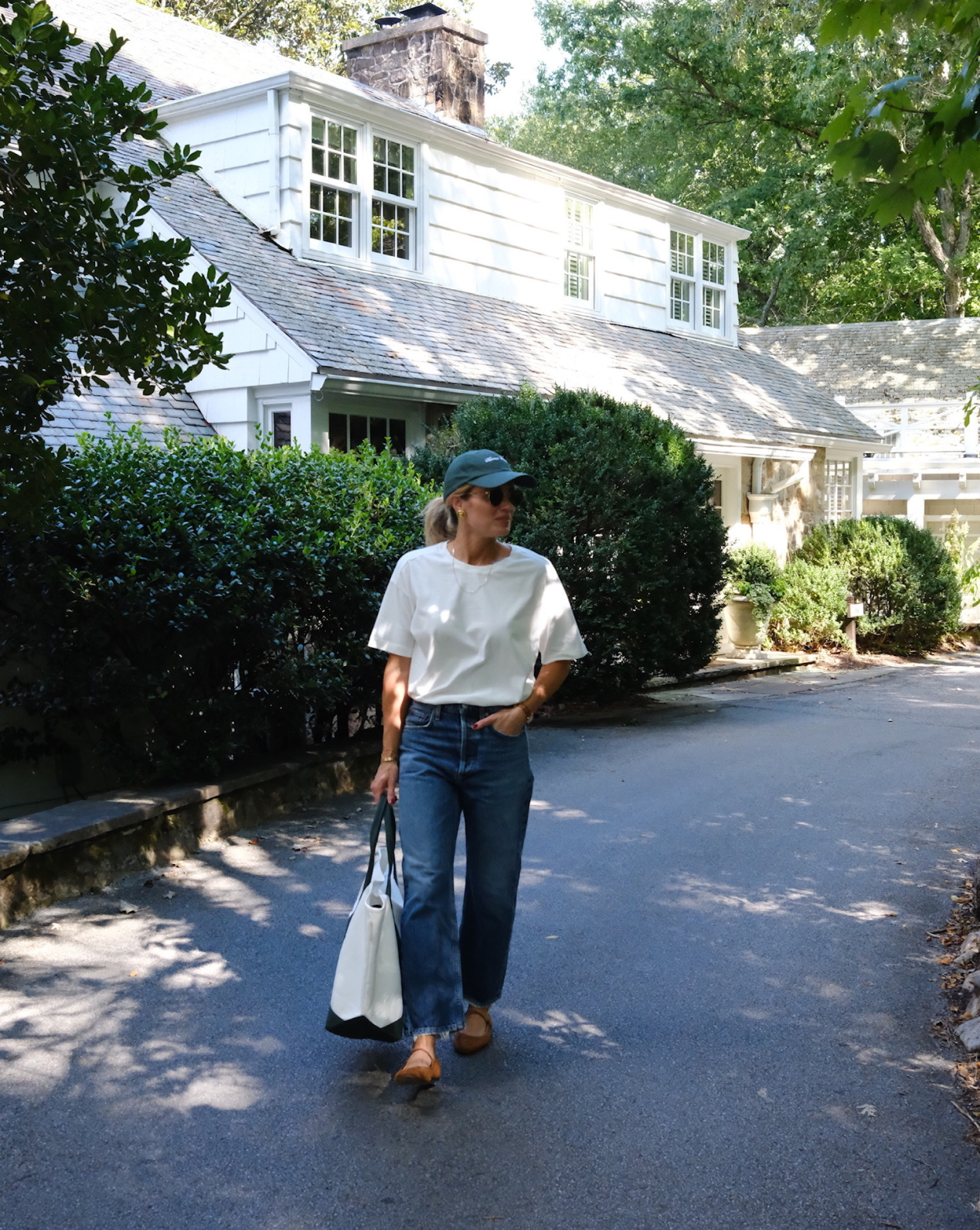 A person in a white t-shirt, blue jeans, green cap, and brown shoes walks on a shaded driveway, carrying the Canvas Laurel Tote from Studio McGee, with a white house and green bushes in the background, by McGee & Co.