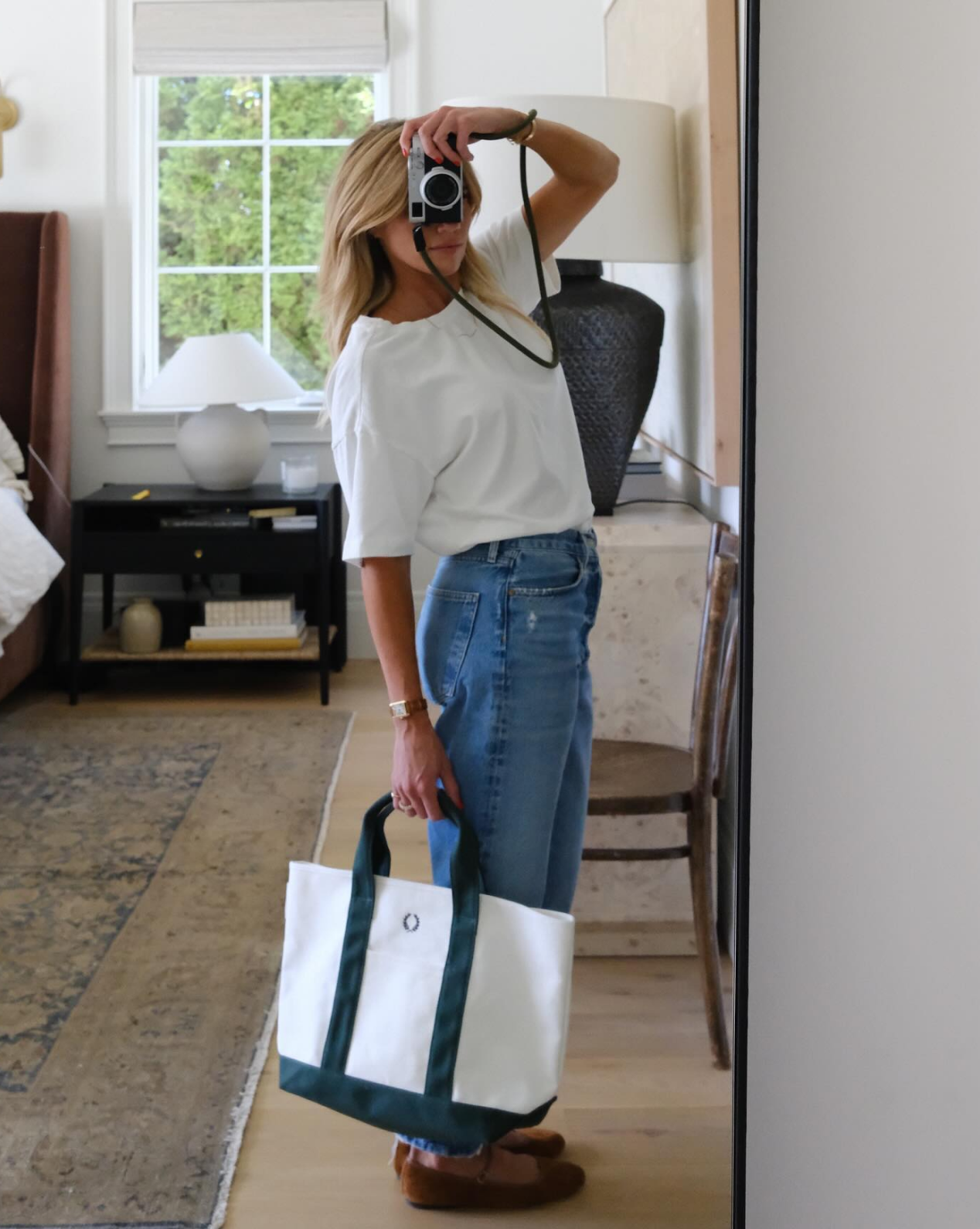A woman stands in a Studio McGee-inspired bedroom taking a mirror selfie. She wears a white shirt, blue jeans, brown shoes, and carries the Canvas Laurel Tote. The room features a bed, rug, chair, and window, by McGee & Co.