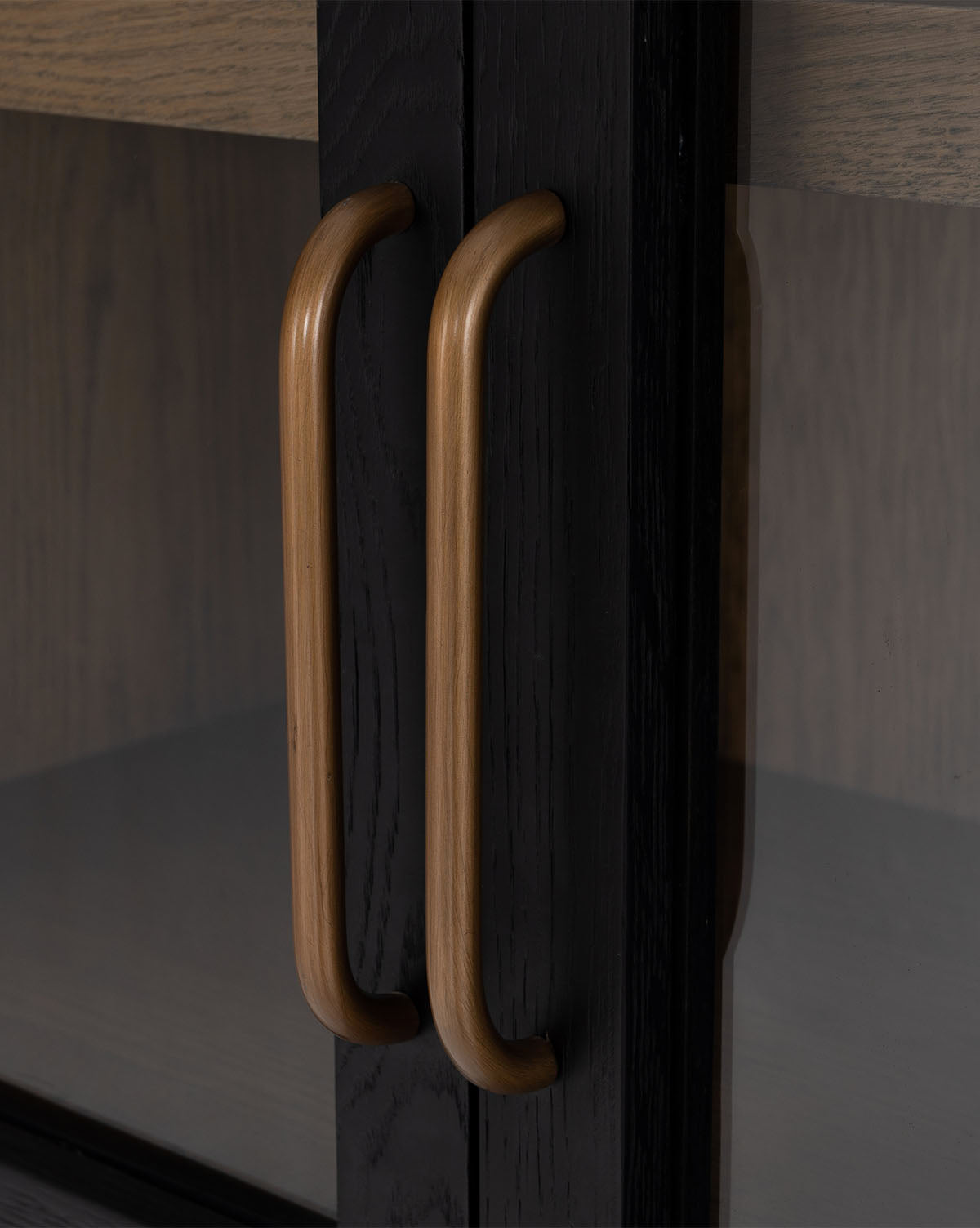 Close-up of two smooth, rounded wooden handles on the glass doors of the Providence Cabinet, revealing interior shelving behind its dark oak finish by McGee & Co.