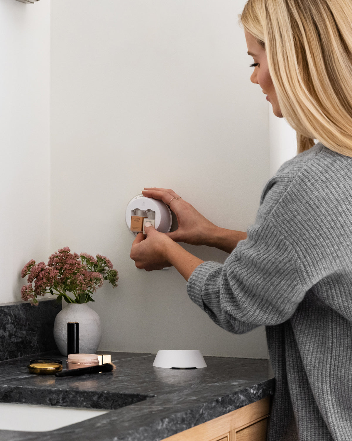 A woman with long blonde hair in a gray sweater places a Pura x Studio McGee Neroli Basil Home Fragrance Refill on a bathroom wall above a dark counter with makeup and pink flowers, filling the space with an herbal citrus scent.