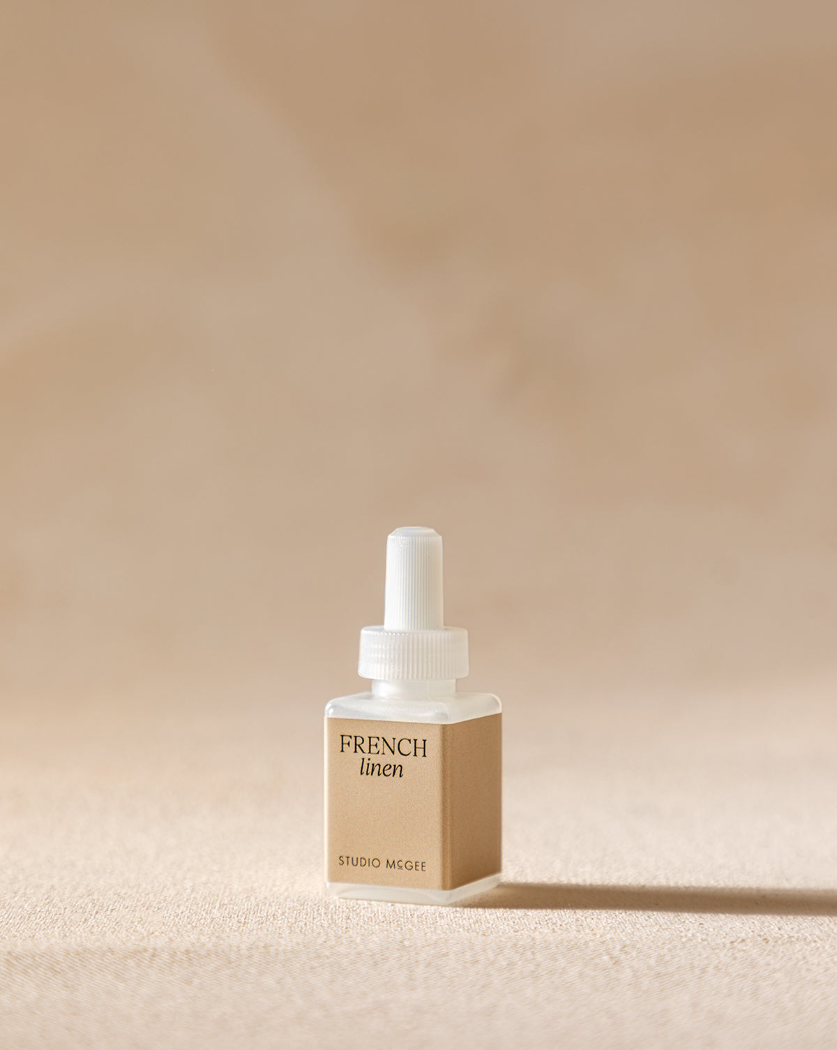 A small, square Pura x Studio McGee French Linen Home Fragrance Refill stands upright on a beige surface, featuring a white cap and dropper against a softly blurred neutral background, suggesting its fresh citrus scent.