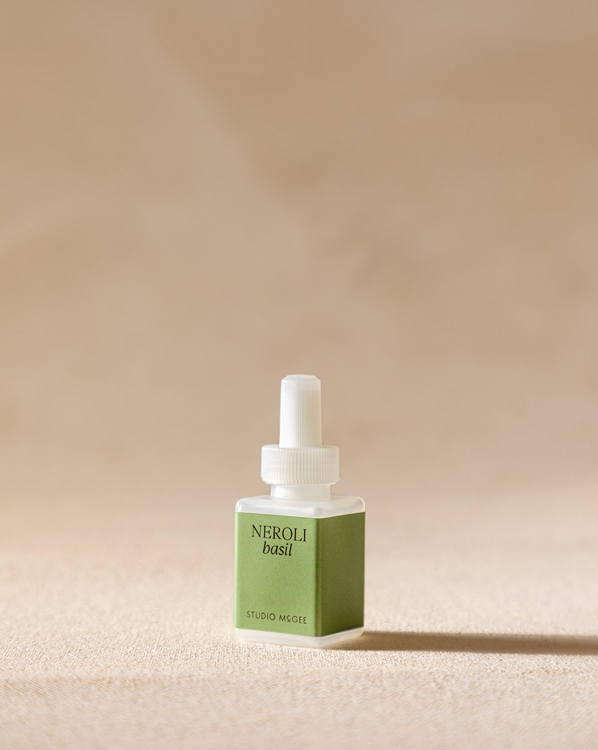 A small square bottle of Pura x Studio McGee Neroli Basil Home Fragrance Refill, with a green label and white cap, rests on a beige surface, evoking an aromatic blend of herbal and citrus notes.