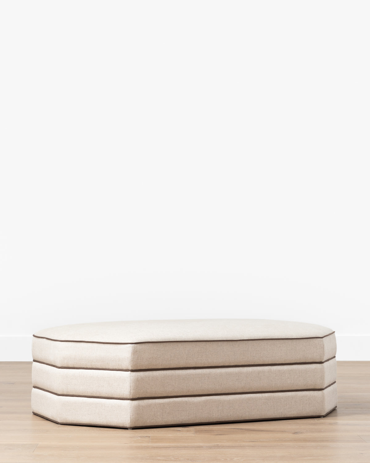 A Makerspalm Rafferty Ottoman in beige performance linen with three stacked cushions rests on a light wood floor against a plain white wall, McGee & Co.