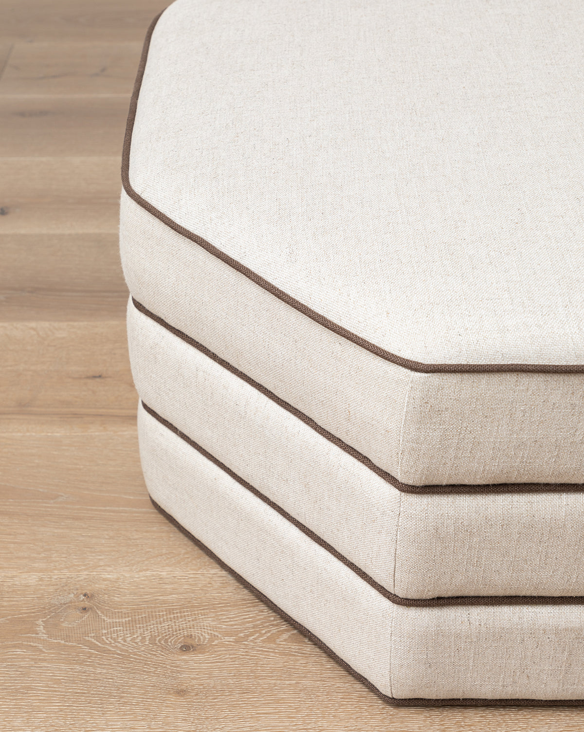 A stack of three beige Rafferty Ottomans by Makerspalm, hexagonal with brown piping and performance linen upholstery, arranged on a wooden floor, McGee & Co.