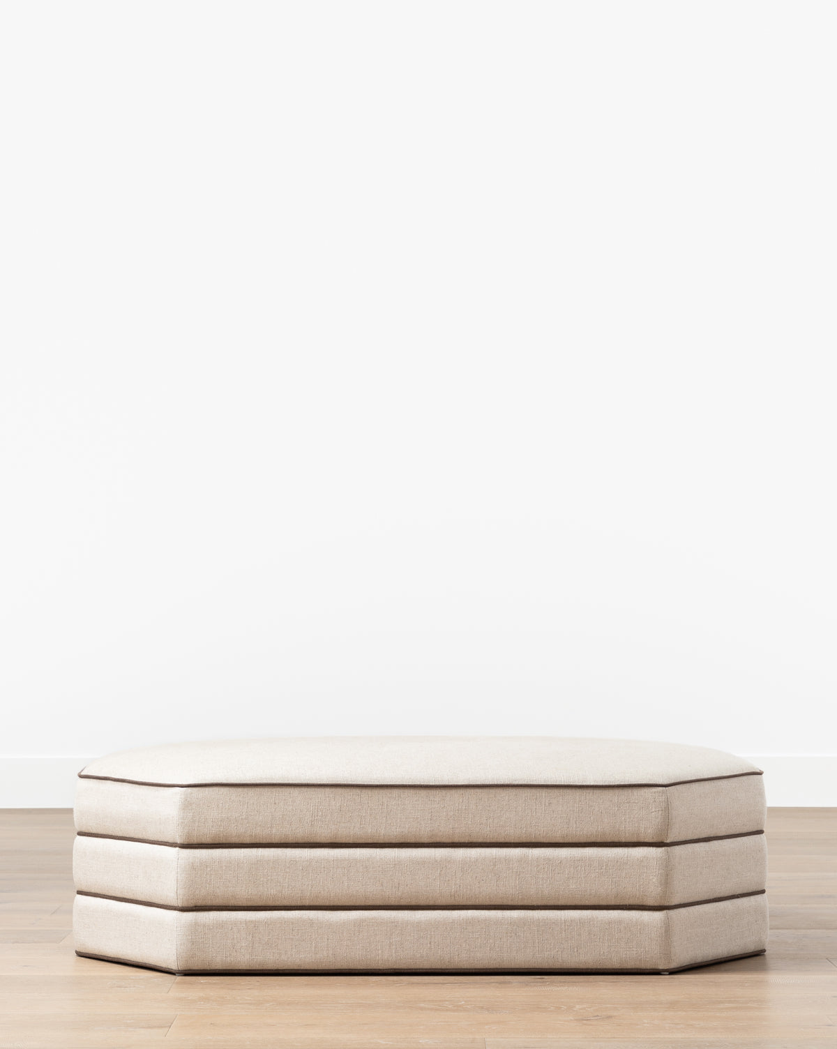The Makerspalm Rafferty Ottoman, a low rectangular piece in beige performance linen with three stacked cushions, is placed on a light wood floor by a plain white wall—McGee & Co.