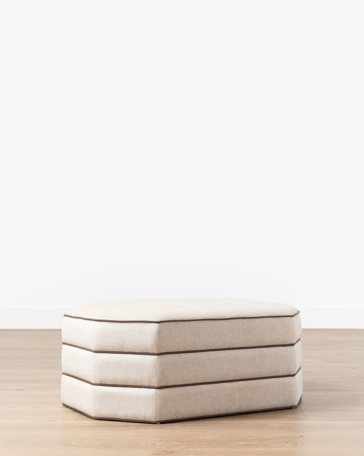 The Makerspalm Rafferty Ottoman, upholstered in beige performance linen with three stacked cushion layers, stands on a light wooden floor against a plain white wall. McGee & Co.