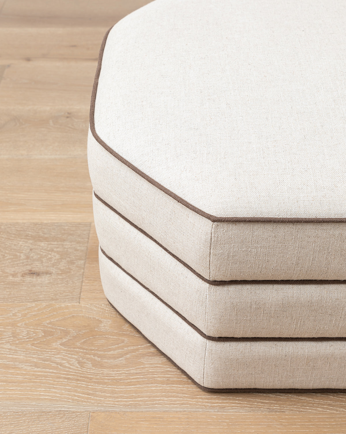 Three beige, octagonal Makerspalm Rafferty Ottoman cushions with dark brown piping in performance linen are stacked on a light wooden floor, McGee & Co.