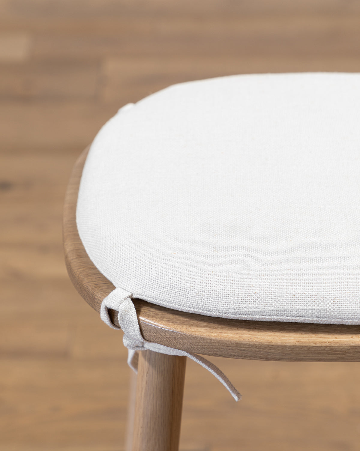 Close-up of a linen cushion tied to the Ralph Oak Counter Stool, with a blurred wooden floor in the background near the kitchen counter, by McGee & Co.
