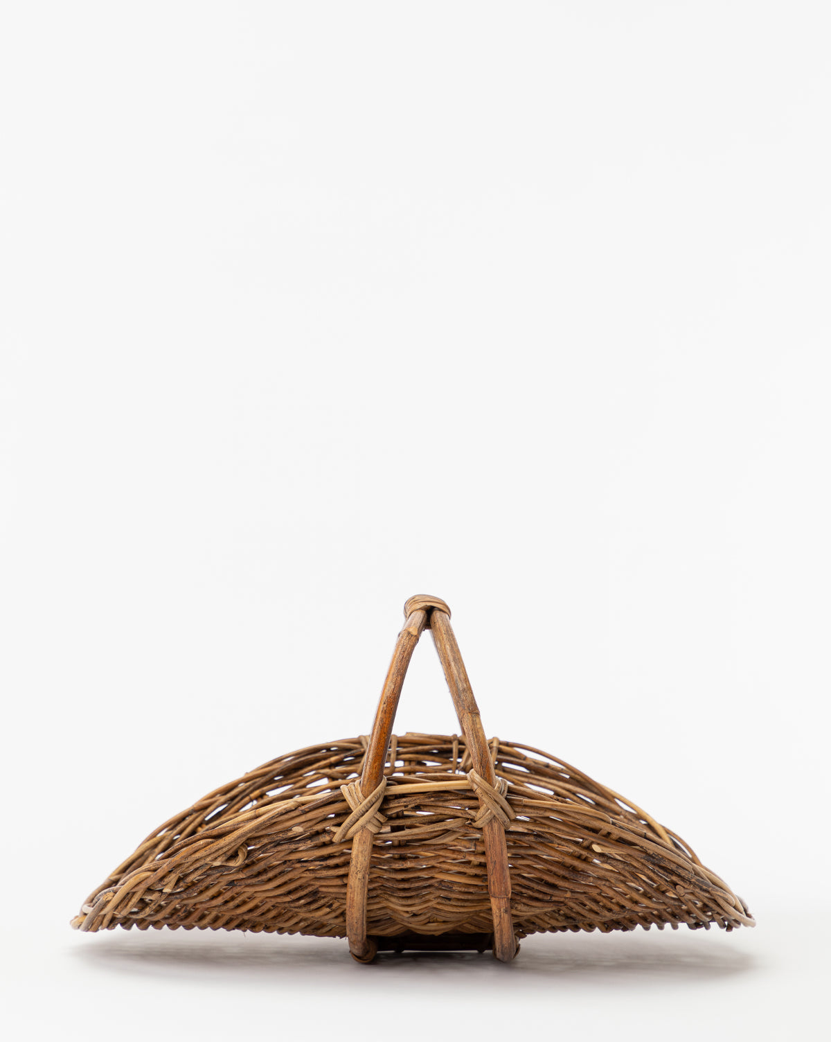 The Rattan Gardening Tray, featuring a woven wicker design and curved wooden handle, sits upright on a plain white background. Its interlaced fibers and arched shape create an organic, rustic look.