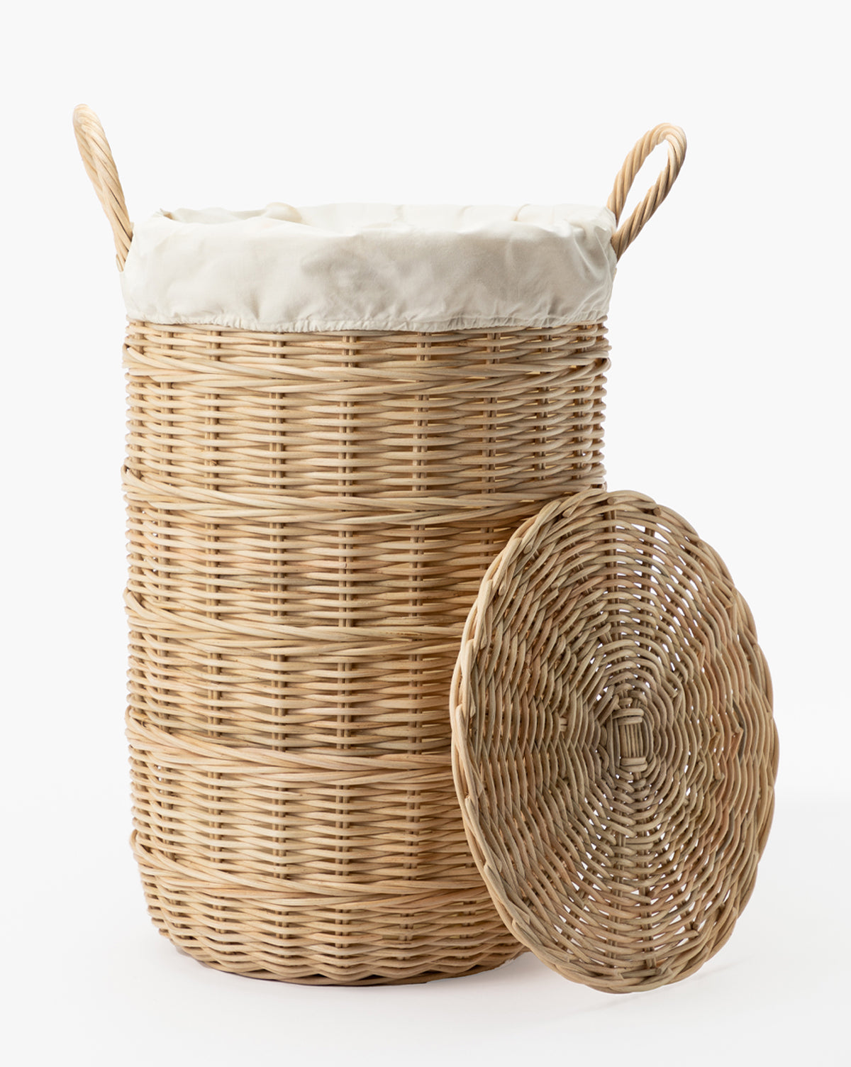 The Rattan Lidded Laundry Bin features a round woven design, two handles, a matching wicker lid, and a white cloth lining. Ideal for stylish laundry rooms. Lid is shown propped in front. Background is plain white.