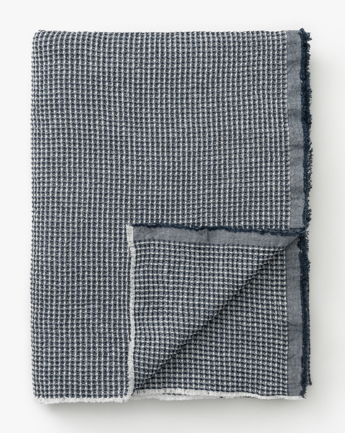 The Rebecca Throw is a folded navy cotton blanket with a waffle weave and grid-like texture, neatly turned to reveal the reverse. It features frayed edges for added style.